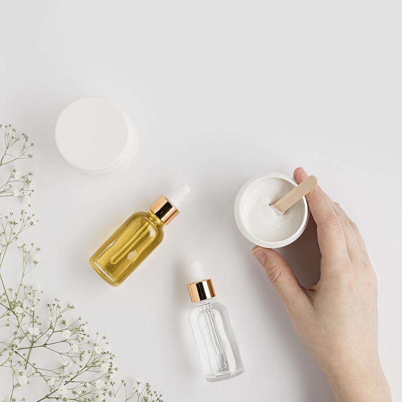 Kosmetiikkatuotteiden asetelma valkoisella pinnalla: pipettipulloja, voidepurkkeja ja valkoisia kukkia.